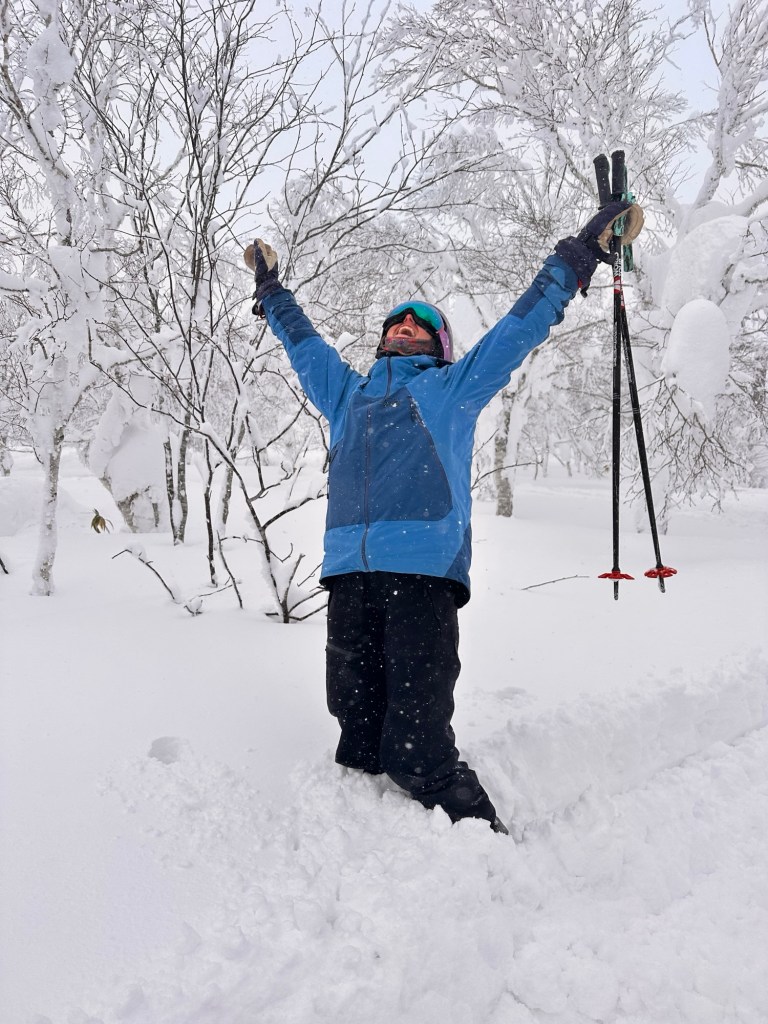 A person stands joyfully in deep snow, surrounded by snow-covered trees, raising their arms in excitement.